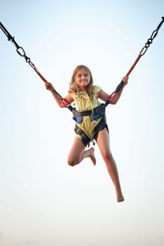 Bungee jumping at trampoline. Little girl bouncing on bungee jumping in amusement park on summer vacations. Kid flying over blue sky while jumping on trampoline. Super girl