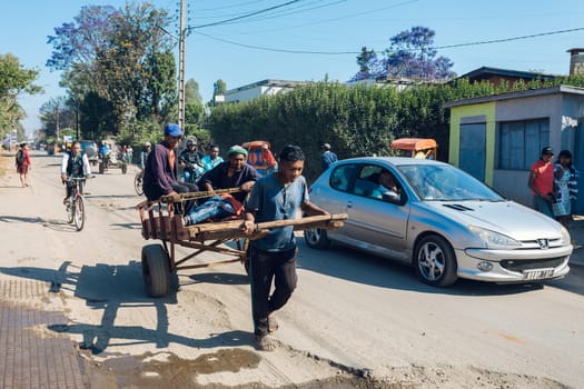 Antsirabe, Madagascar - November 2nd 2022: Rickshaw runner dashes through the crowded streets with small wooden cart. A handheld rickshaw driven by a running person is traditional mode of transport.