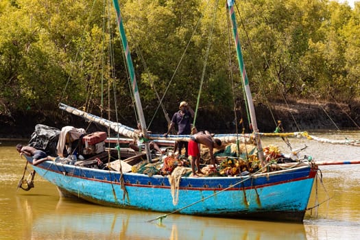 MORONDAVA, MADAGASCAR - AUGUST 28 2023: Two men repair a fishing net on a blue boat moored to the riverbank in Morondava. The boat is equipped with two masts and other fishing gear.