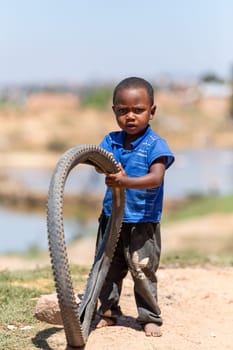 Antananarivo, Madagascar - November 27th 2022: A young boy plays with an old bicycle tire while standing on a dirt road.