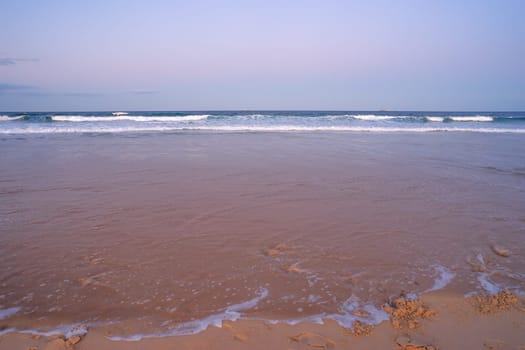 The foreshore covered by water as the tide rises on a sandy surf beach showing surf, waves, breaking water,and ships on the distant horizon at dusk