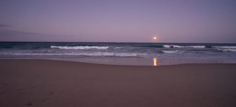 A blue moon, full moon, rising over the Pacific Ocean, Sunshine Coast, Eastern Australia at the end of August, 2023, with ships on the horizon and surf waves crashing onto a sandy beach.