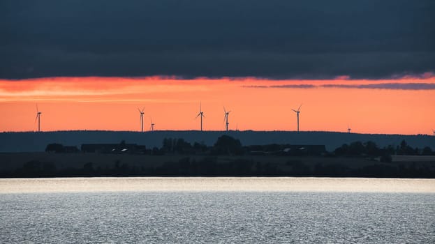 Wind turbine, renewable energy, on a lake at sunset. Clean electricity for climate change. Cloudy sky with orange sunshine in Scandinavia.