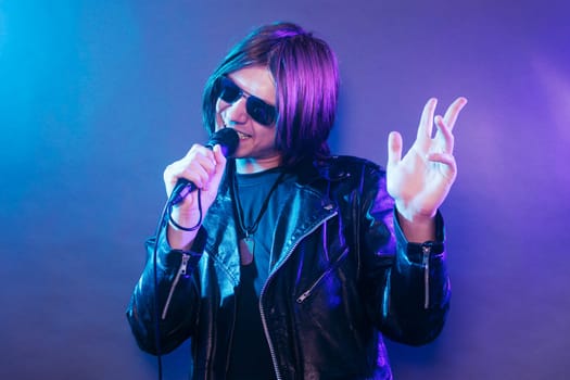The young rock star in black leather is singing in microphone on blue purple background.
