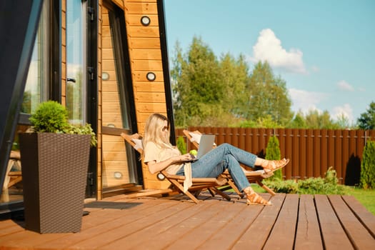 Woman watching movie on laptop while resting in a folding wooden sun lounger on the terrace of log cabin