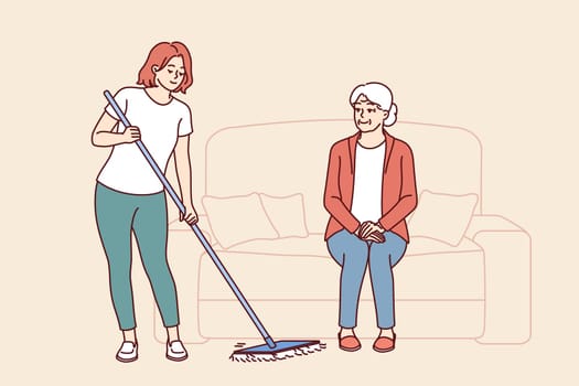 Caretaker from nursing home is mopping floor and taking care of elderly woman sitting on couch. Gray-haired female pensioner with smile watches caretaker cleaning room to maintain cleanliness