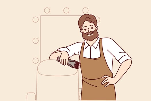 Bearded man hairdresser works in barbershop and smiles, standing near chair for client and mirror. Successful stylist in apron posing indoors at barbershop and holding trimmer in hand
