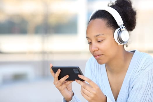 Black woman wearing headphone watching video on horizontal phone in the street
