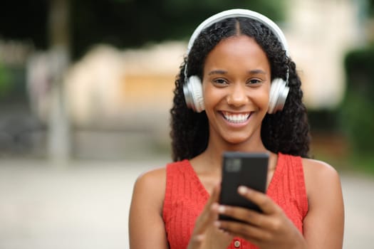 Front view portrait of a happy black woman wearing headphone holding phone looking at camera