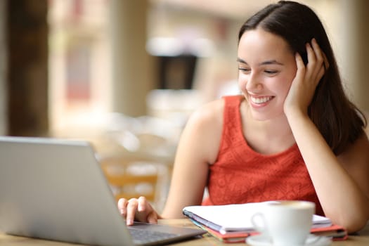 Happy student elearning using laptop in a bar terrace