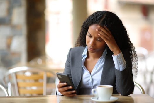 Worried black businesswoman checking bad news on phone in a bar terrace