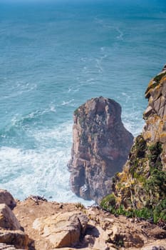 Scenic view of steep rock in blue lagoon near beach seashore coastline surrounded by stormy waves, sheer mountains cliffs covered with green grass. Summer, holiday, travelling, seascape, tourism.
