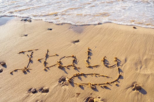 Close-up of inscription word Italy written on wet sand of beach seashore coastline after tide near sea waves on sunny day. Summertime, holiday, vacation, travelling, seascape, tourism, handwriting.