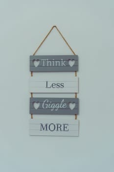 Composition of wooden plaques boards with inscription words think less giggle more, holes in shape of hearts joined with rope hanging on wall on grey background. Wall decoration, placard. Isolated.