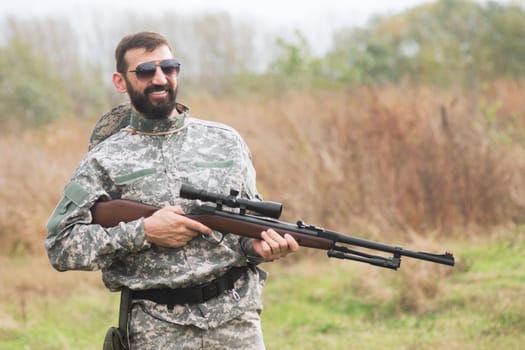 The smiling army soldier in military uniform is holding a shotgun or airgun weapon outdoors.