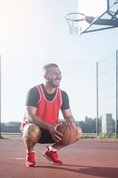The young smiling basketball player with basketball ball is squatting on playground.