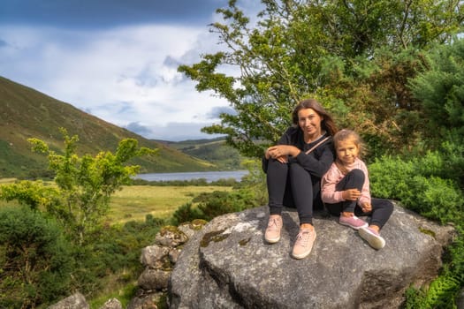 Mother and daughter, family, sitting on a large rock and smiling. Lake Lough Dan and mountain valley in background, Wicklow Mountains, Ireland