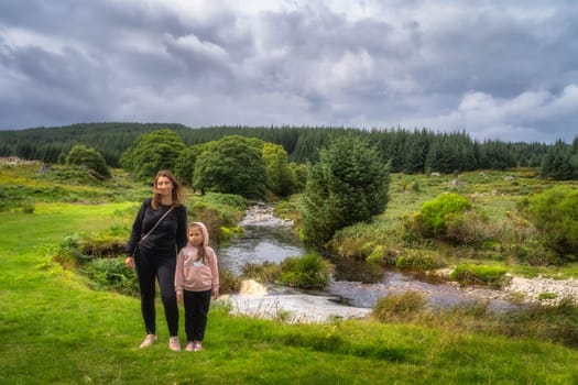 Mother and daughter standing on grass, with winding river and green forest in background. Family hike in Lough Dan, Wicklow Mountains, Ireland