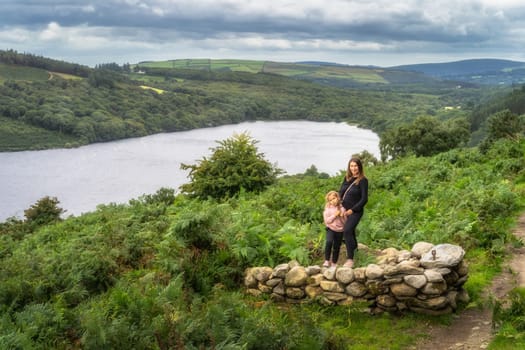 Mother and daughter standing on rocks, with lake, Lough Dan, forest and fields in background. Family hiking in Wicklow Mountains, Ireland