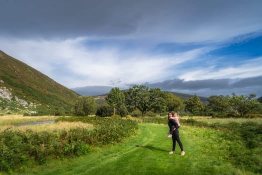 Young girl piggyback riding on her mum on a country road with river and green forest in background. Family hike, Lough Dan, Wicklow Mountains, Ireland