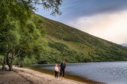 Mother and daughter standing on the beach, with mountain and forest in background. Family hike in Lough Dan, Wicklow Mountains, Ireland