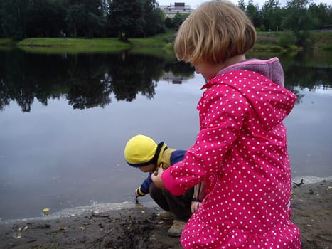 Brother and sister are walking near the river. A boy and a girl are dressed in jeans and autumn clothes. Early summer. Talk and play by the water. Pink cloak in white polka dots, yellow jacket.