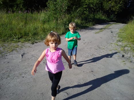 Blond-haired children walk on a dirt road in the summer. Green T-shirt. Girl in a pink blouse with frill. Brother and sister rush home. Hiking in nature in sunny weather. Sweeping movements