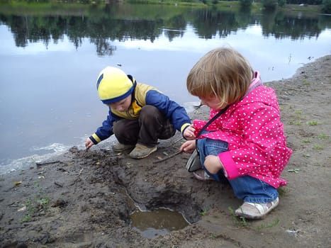 Brother and sister are walking near the river. A boy and a girl are dressed in jeans and autumn clothes. Early summer. Talk and play by the water. Pink cloak in white polka dots, yellow jacket.