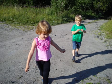 Blond-haired children walk on a dirt road in the summer. Green T-shirt. Girl in a pink blouse with frill. Brother and sister rush home. Hiking in nature in sunny weather. Sweeping movements