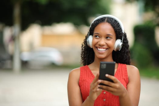 Front view portrait of a happy black woman wearing headphones and holding phone looking at side listening music