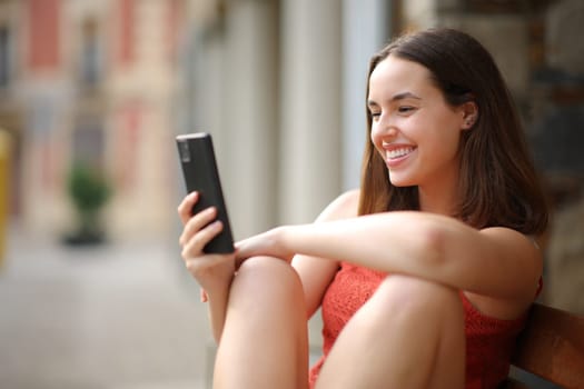 Happy woman sitting on a bench in the street using smart phone