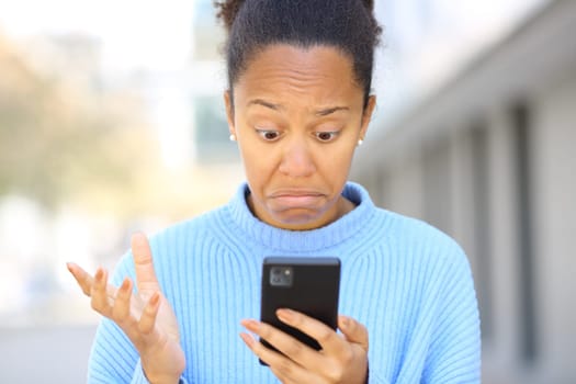 Front view portrait of a perplexed black woman checking cell phone in the street