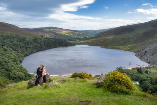 Mother and daughter, family, sitting on a large rock on the edge of cliff. Lough Tay, called Guiness Lake in Wicklow Mountains, Ireland