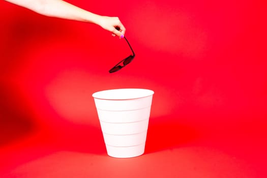 Hand throws sunglasses into a trash bin with package on red background
