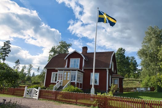 a typical red and white swedish house in smalland. White garden gate, brown fence. Flagpole with sweden flag. green lawn in garden. Trees in the background. Blue sky with small white clouds