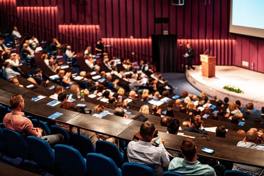 Business and entrepreneurship symposium. Female speaker giving a talk at business meeting. Audience in conference hall. Rear view of unrecognized participant in audience.
