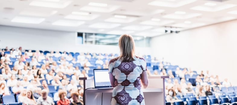 Female speaker giving a talk on corporate business conference. Unrecognizable people in audience at conference hall. Business and Entrepreneurship event.
