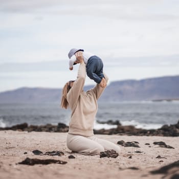 Mother enjoying winter vacations holding, playing and lifting his infant baby boy son high in the air on sandy beach on Lanzarote island, Spain. Family travel and vacations concept.