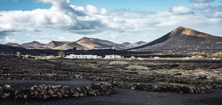 Traditional white houses in black volcanic landscape of La Geria wine growing region with view of Timanfaya National Park in Lanzarote. Touristic attraction in Lanzarote island, Canary Islands, Spain