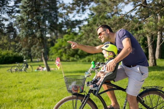 Look over there. Active family day in nature. Father and son ride bike through city park on unny summer day. A cute boy is sitting in front bicycle chair while father rides bicycle. Father son bonding