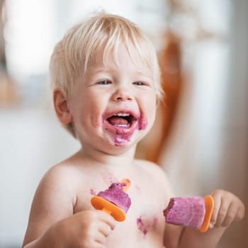 Happy adorable infant baby boy child smiling while eating two frozen fruit popsicle ice creams in simmer