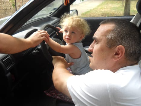 A small child is driving a car. A middle-aged father supports him with his hands. The boy plays in the driver of the car. Dad and son look away and listen to someone outside the vehicle.