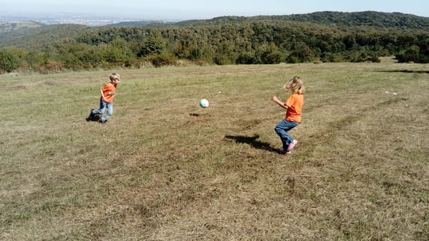 A boy and a girl with blond hair, dressed in orange t-shirts and classic jeans, play ball in a mountain meadow. Early warm autumn. Kids are playing. In the background the horizon and vegetation.