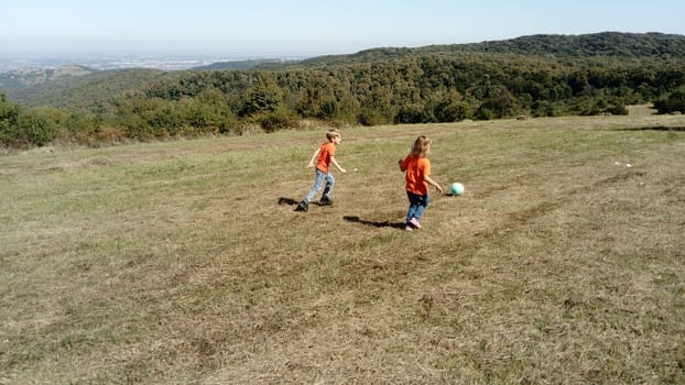 A boy and a girl with blond hair, dressed in orange t-shirts and classic jeans, play ball in a mountain meadow. Early warm autumn. In the background, the horizon and vegetation