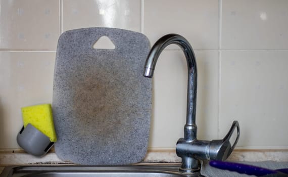 Water faucet and gray cutting board in the background. Dirty cutting board near the sink, unwashed dishes after cooking dinner. Daily chores