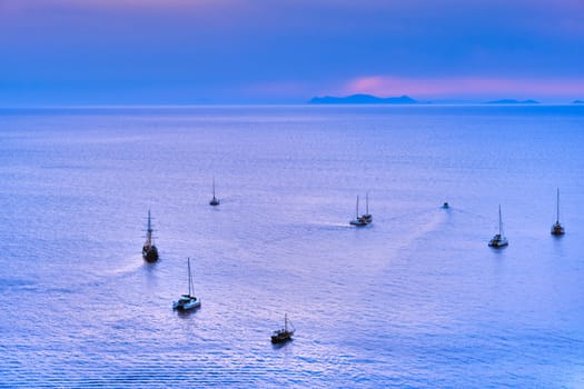 Aerial view of numerous sailboat and yachts go to sea for sunset or sunrise. Calm sea waters, hazy distant sun rays, blue sky and clouds, seascape, cloudscape. Adventure, summer vacations, tourist attraction.