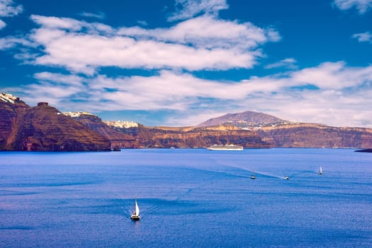 Panorama of Santorini island, Greece. Rough cliffs of volcanic island, sunny day, caldera bay, blue sky and clouds, Fira or Thira town on hilltop, sailboats and yachts in harbor. Summer cruise, vacations, travel destination, tourist attraction, sea