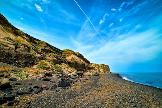 Dramatic landscape of blue sea or ocean waves rolling on rocky volcanic beach on cloudy day. Blue sky, beautiful clouds, black lava pebbles and sand, weathered cliff. Santorini island, Greece.