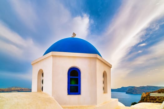 White Greek Orthodox church and its blue dome with cross against dramatic blue sky and clouds, Santorini island, Greece. Beautiful landscape, sea waters, islands, harbor, cliffs, travel destinations in Mediterranean sea cruises, tourist attractions