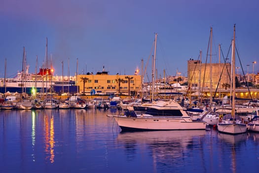 View of port area and bay in Heraklion, capital of Crete island, Greece. Ferries and cruises, fishing boats moored in harbor at sunset. Clear blue sky, deep blue waters, yachts, sailboats. Travel destination of Mediterranean sea, summer vacations.